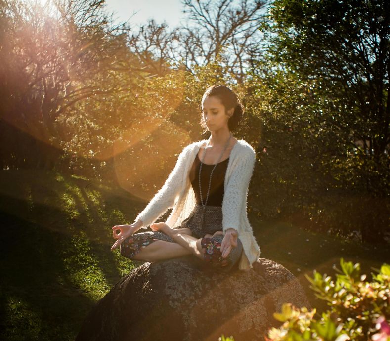 Woman in tranquil meditation pose outdoors, bathed in sunlight, surrounded by lush greenery.