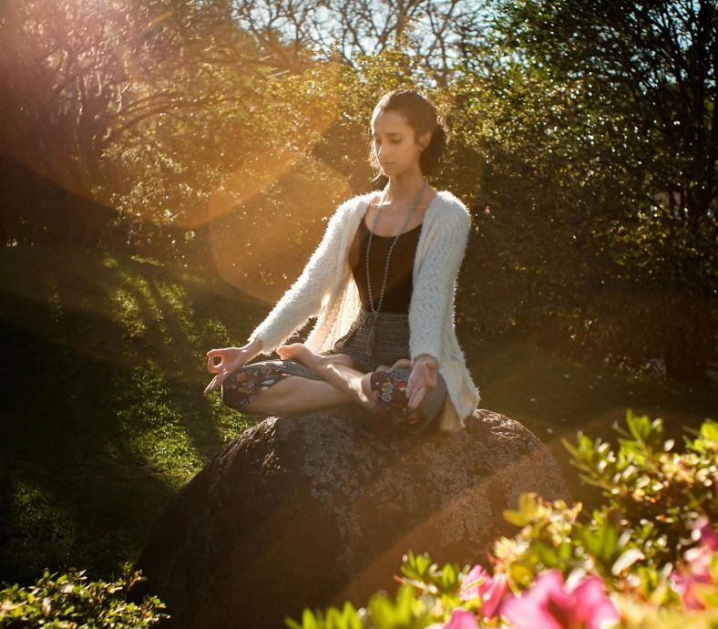 Woman in tranquil meditation pose outdoors, bathed in sunlight, surrounded by lush greenery.